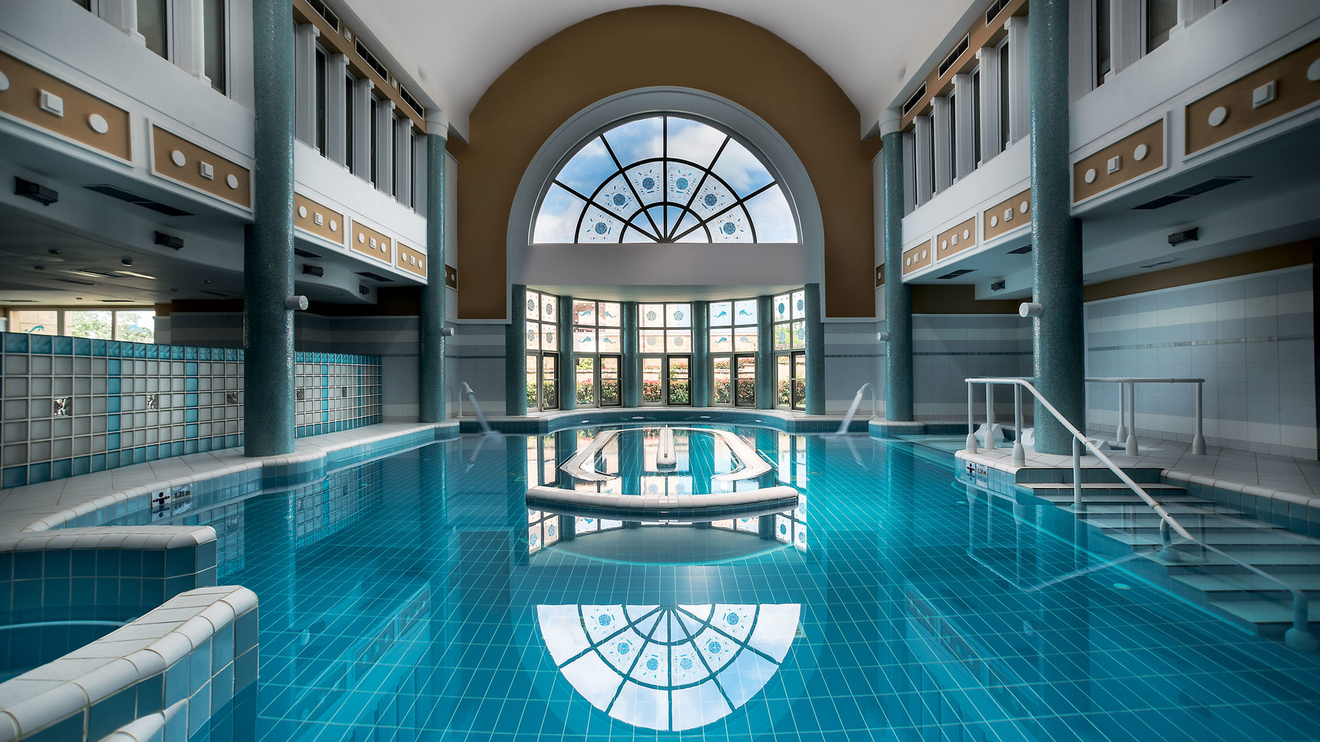 Indoor pool with columns and a tall arched window reflected in the still turquoise water, with steps and handrails