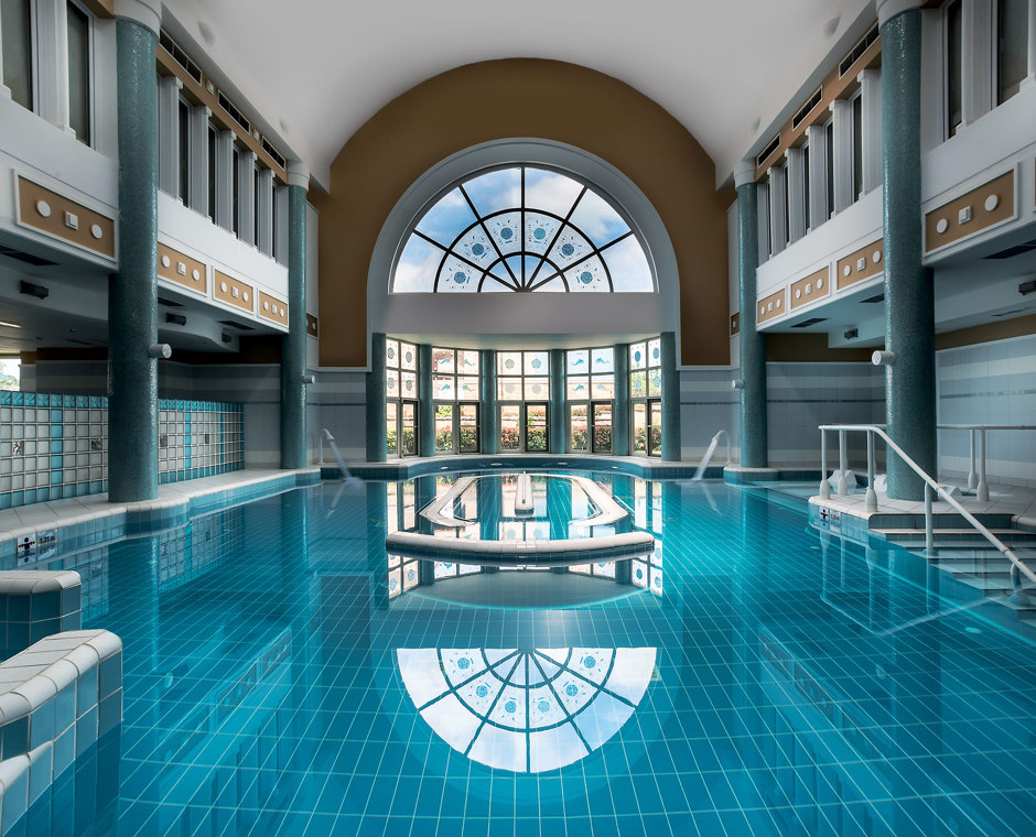 Indoor pool with columns and a tall arched window reflected in the still turquoise water, with steps and handrails