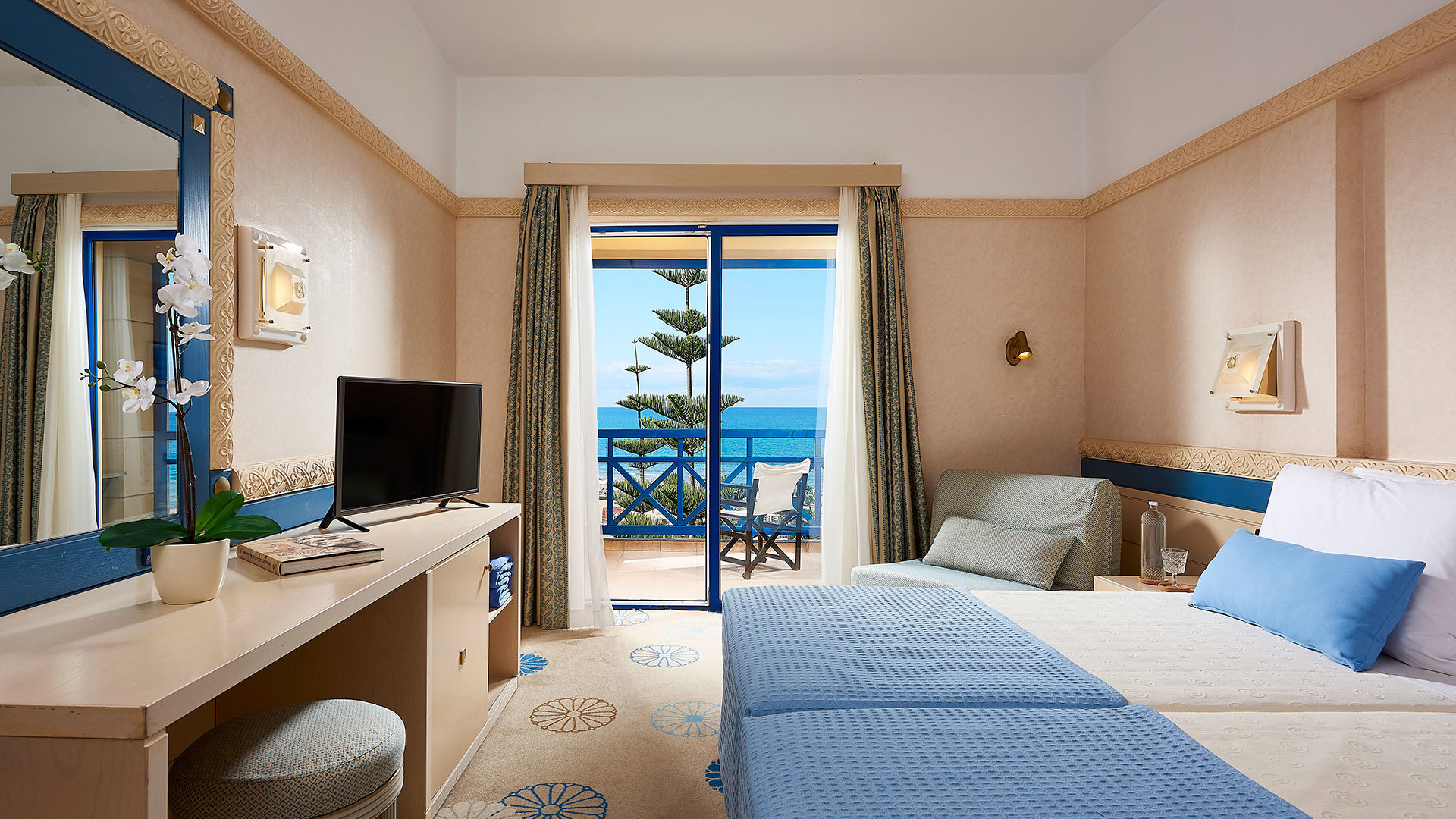Room with two beds in blue covers, desk with TV and mirror, armchair and open balcony door with sea and pine tree view