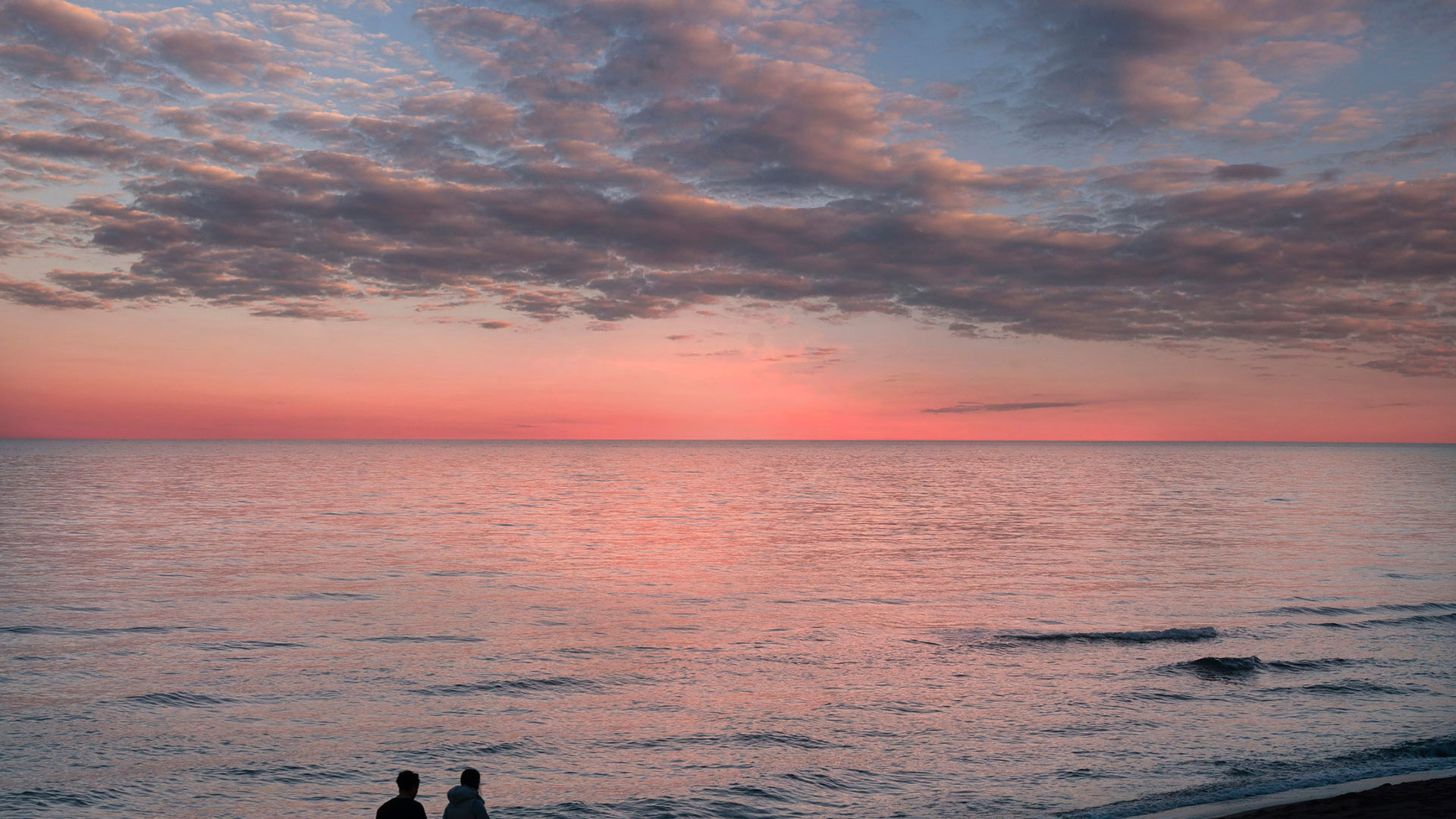 Couple walking along the shore at sunset, under a pink sky with soft clouds over the calm sea