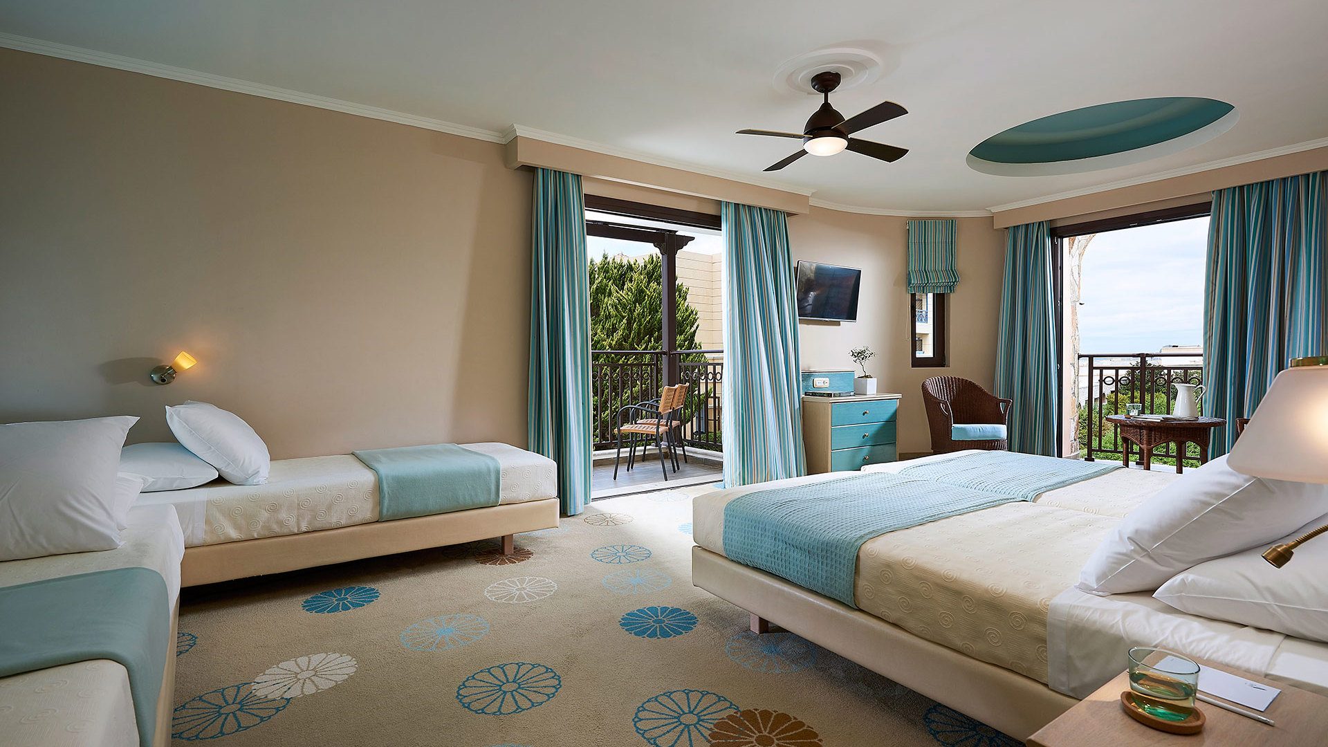 Spacious room with a double bed and two single beds, ceiling fan, turquoise curtains and two balcony doors with sea view