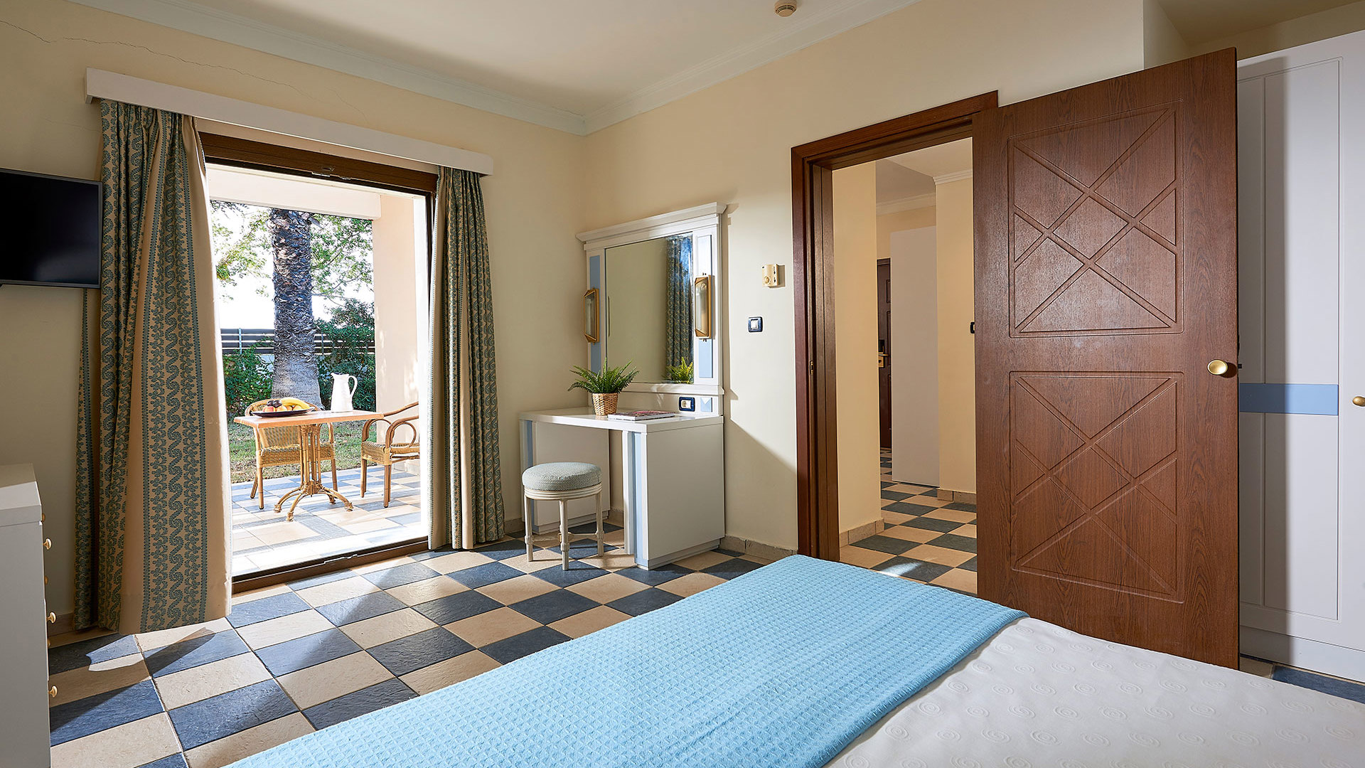 Room with a bed in a turquoise throw, checkered floor, vanity with mirror, wooden door and open doors to a garden terrace