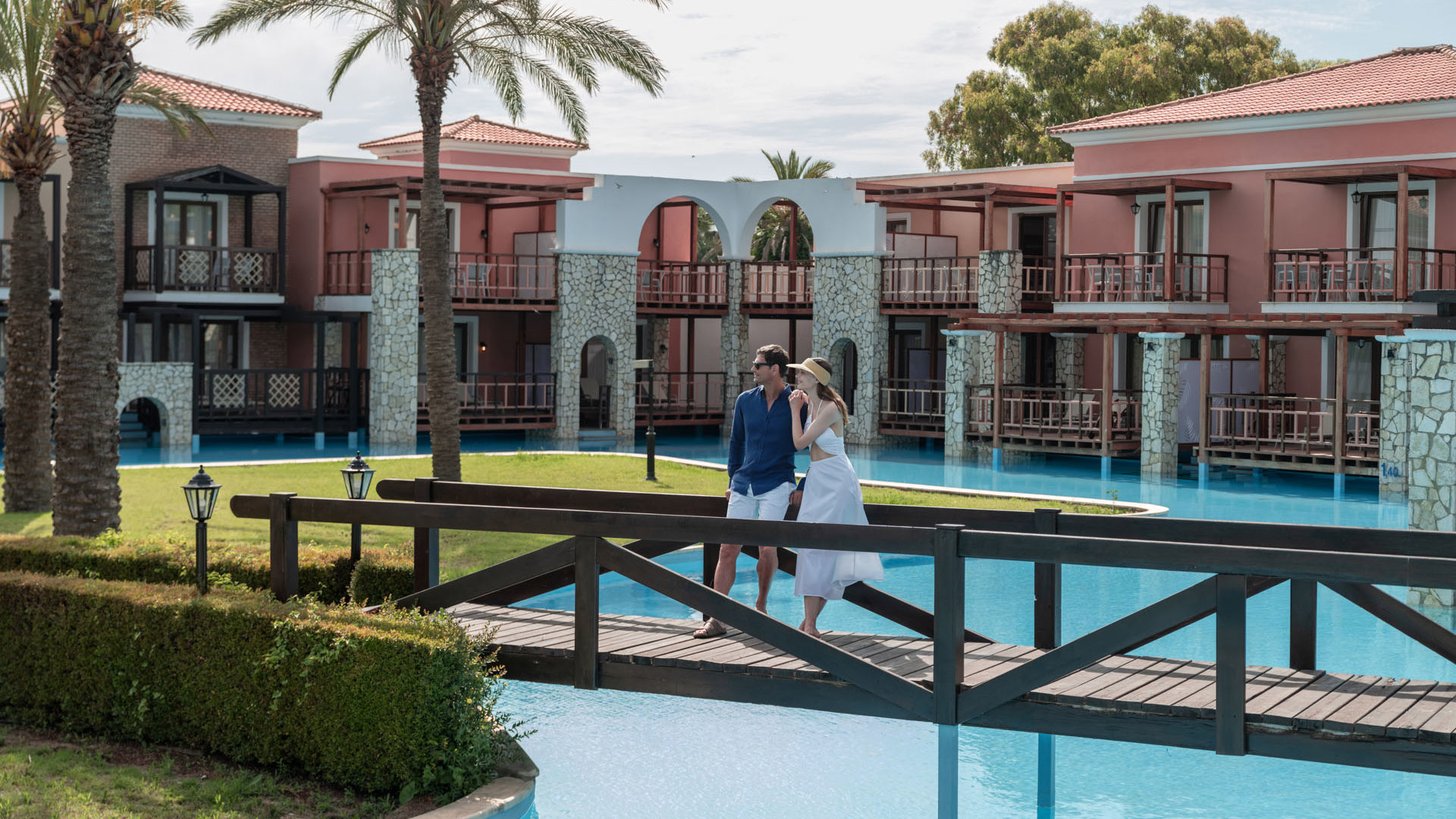 Couple crossing a wooden bridge over the pool, with palm trees and bungalows with balconies at Aldemar Olympian Village
