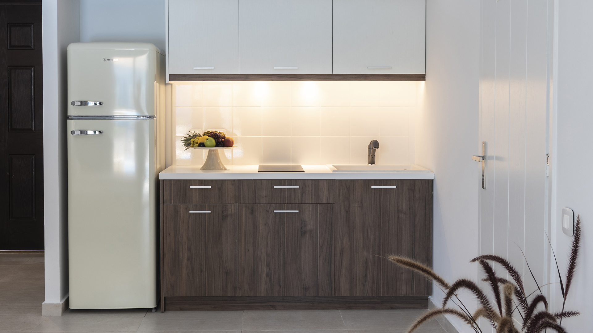 Kitchenette with a retro cream fridge, dark wood cabinets, white upper cupboards, sink, cooktop and a bowl of fruit