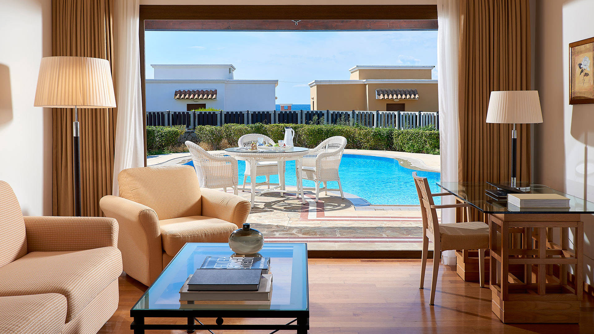Lounge with cream sofa and armchairs, glass coffee table, floor lamps and a large window overlooking sunbeds and a pool