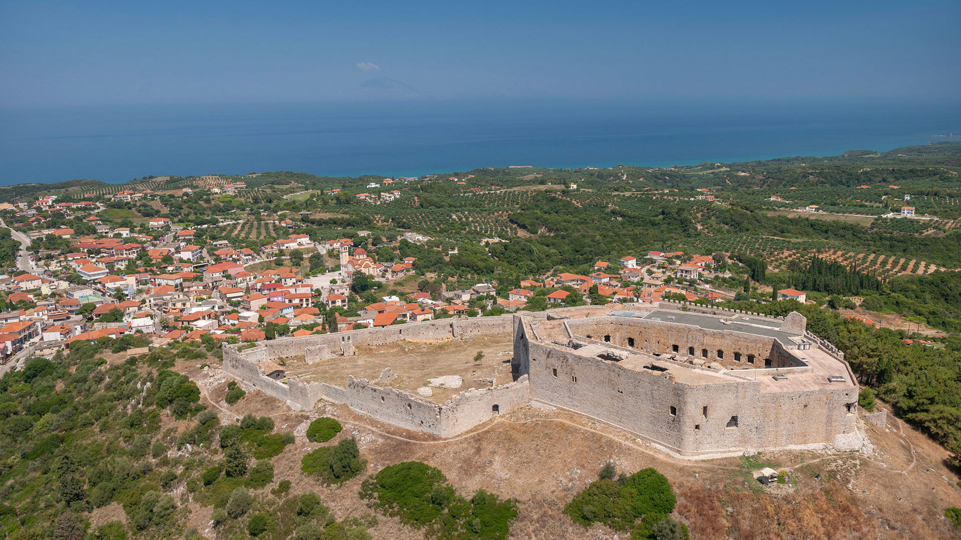 Aerial view of Chlemoutsi Castle on a green hill above the village, with the sea stretching to the horizon