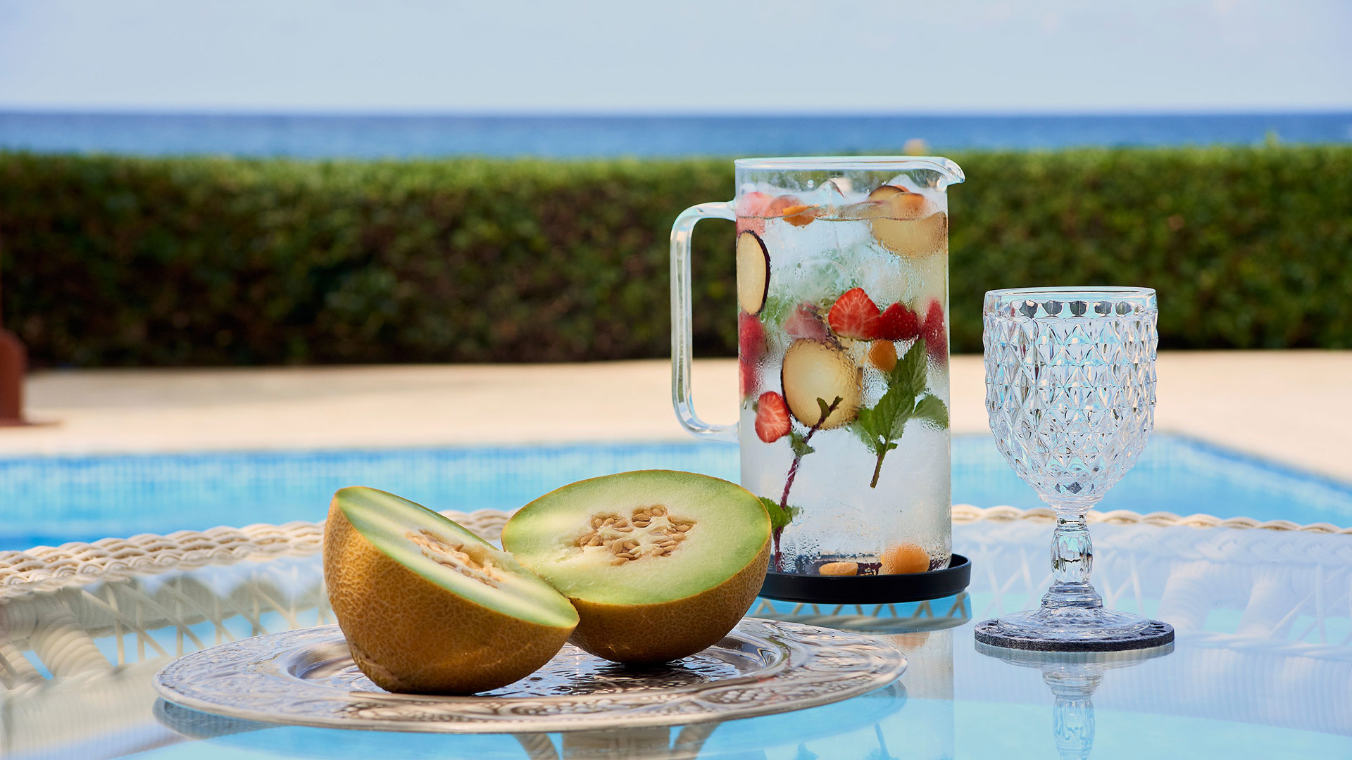 Jug of fruit-infused water with mint, a halved melon and a crystal glass on a table by the pool and sea