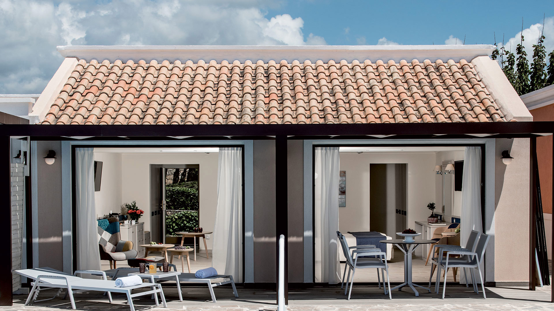 Bungalow with a tiled roof and open glass doors, terrace with sunbeds and chairs beside a blue pool under a cloudy sky
