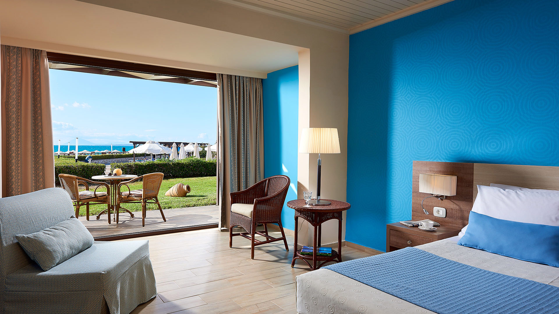 Room with a blue wall, bed, armchairs and open glass doors to a terrace with table and chairs, a lawn and sea view