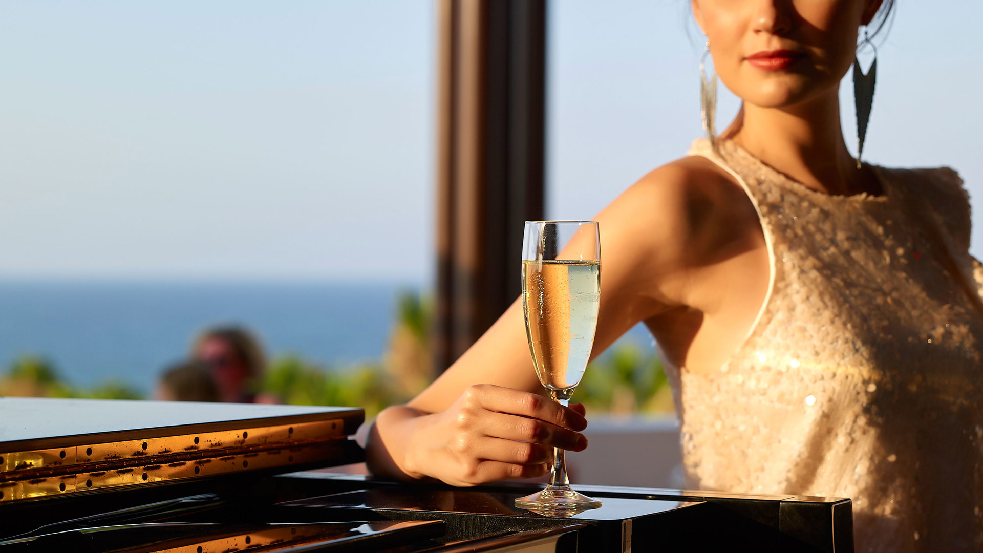 Elegant woman leaning on a black piano with a glass of champagne in hand, the sea glowing in the warm evening light