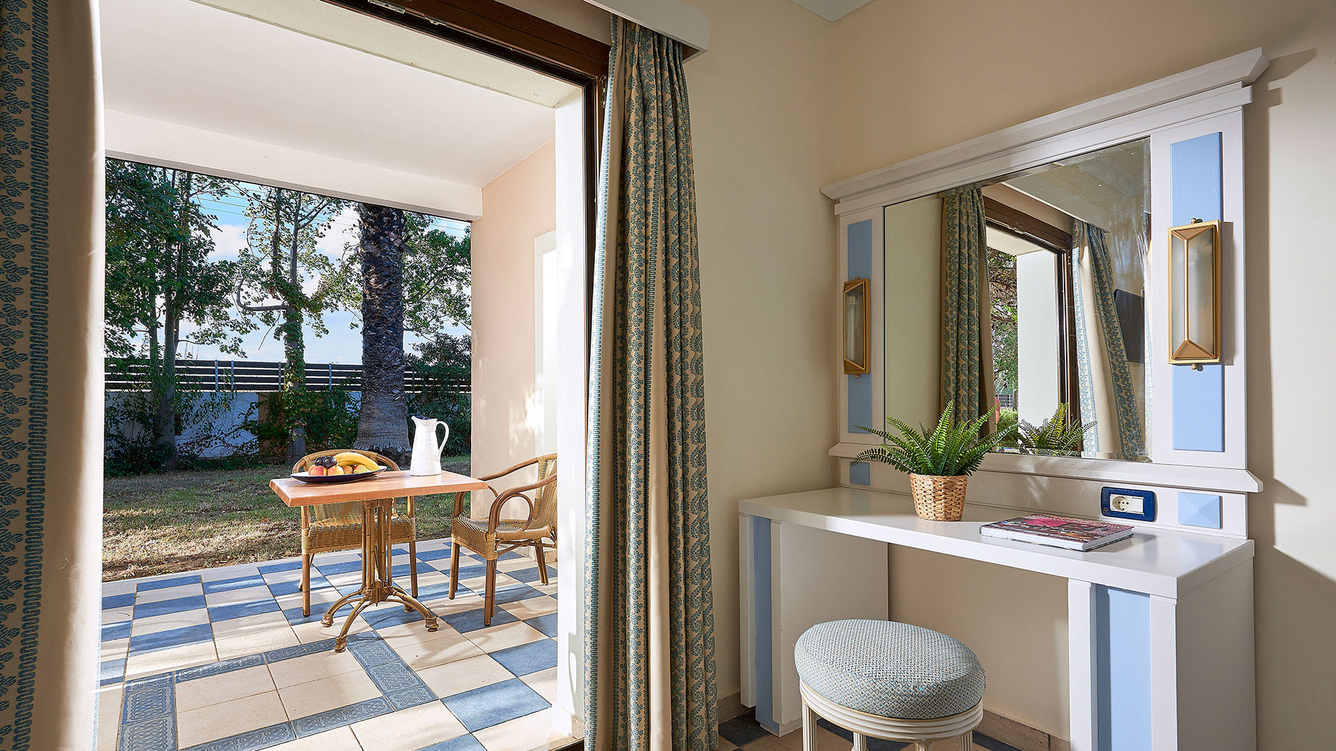 White vanity with a mirror, plant and stool beside open glass doors to a shaded terrace with table, chairs and trees
