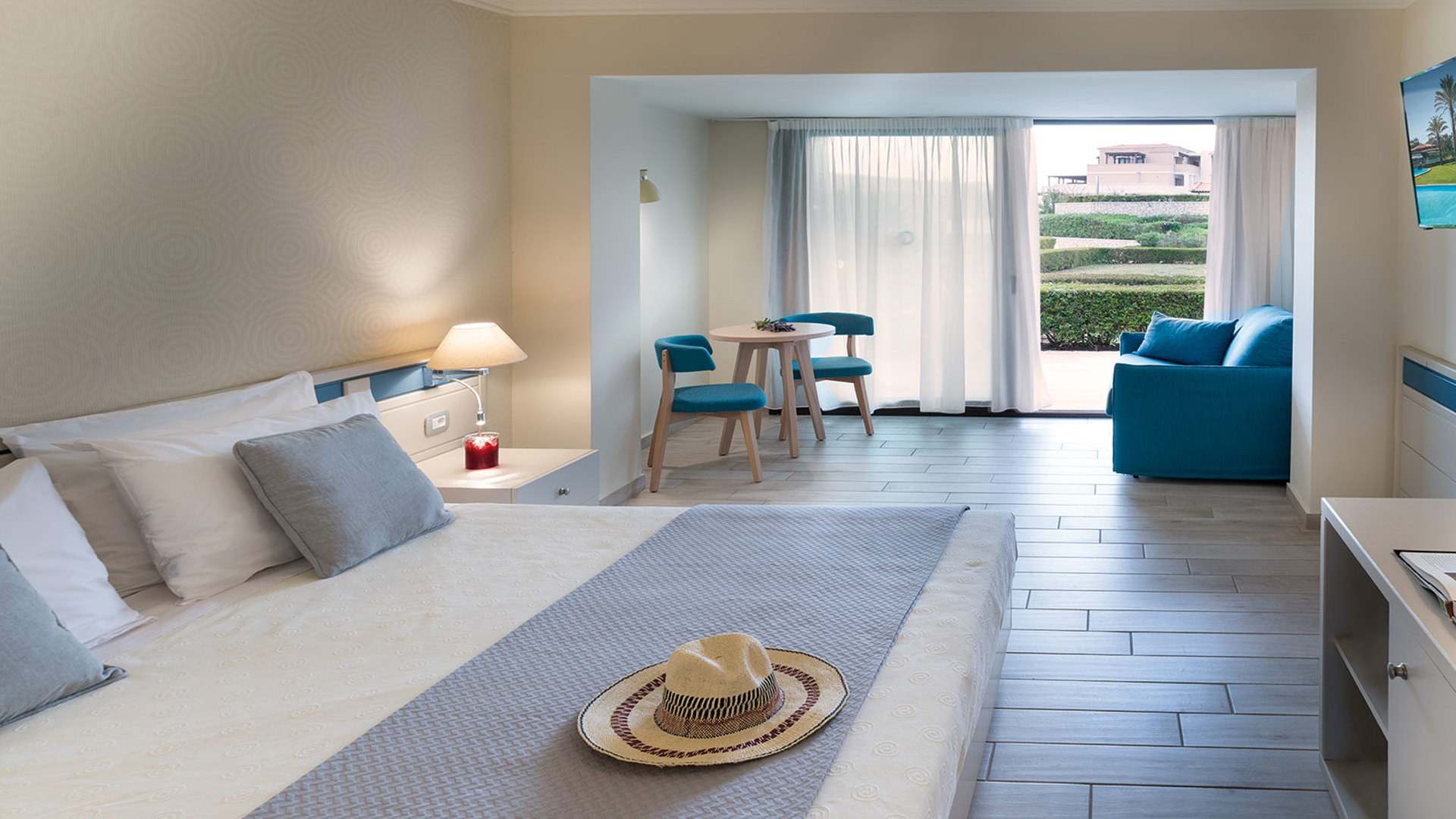 Room with a double bed and straw hat, wall-mounted TV, blue armchair, small round table and balcony door to greenery