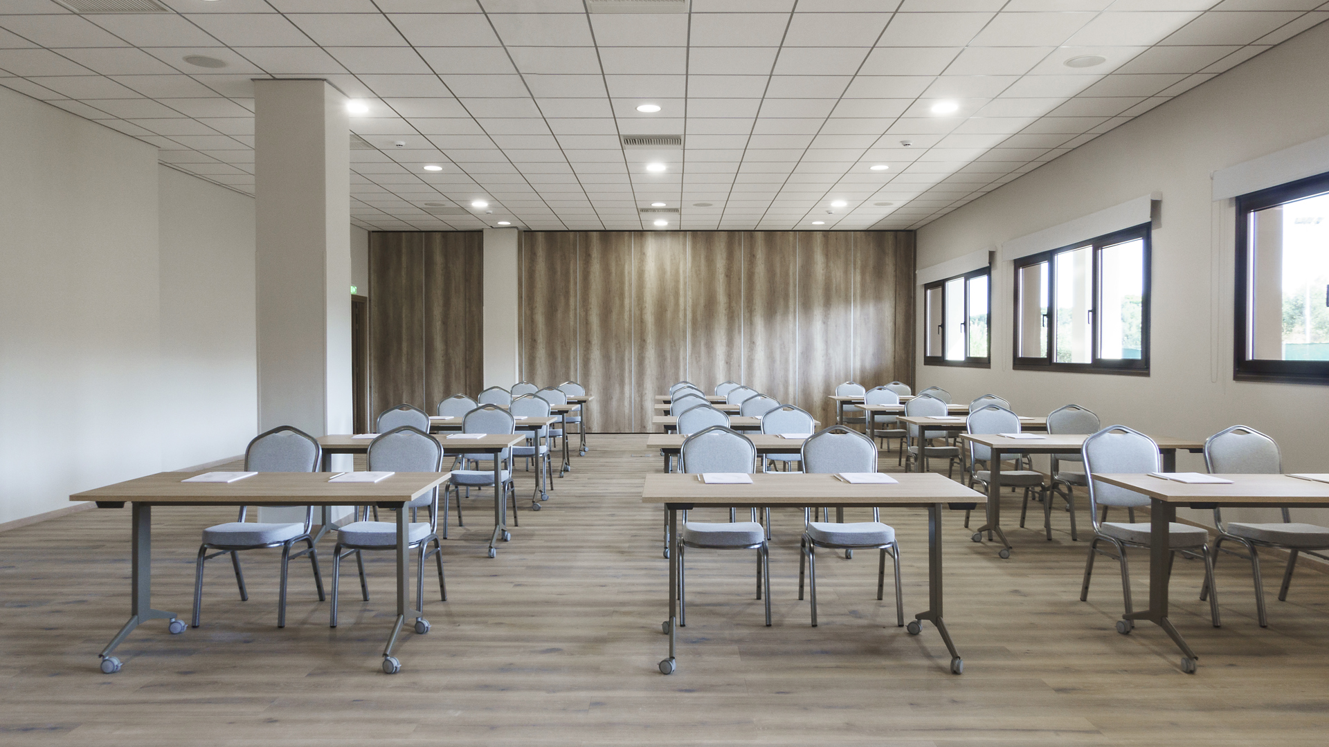 Meeting room in classroom style, with rows of desks and grey chairs, a wood-panelled wall and windows
