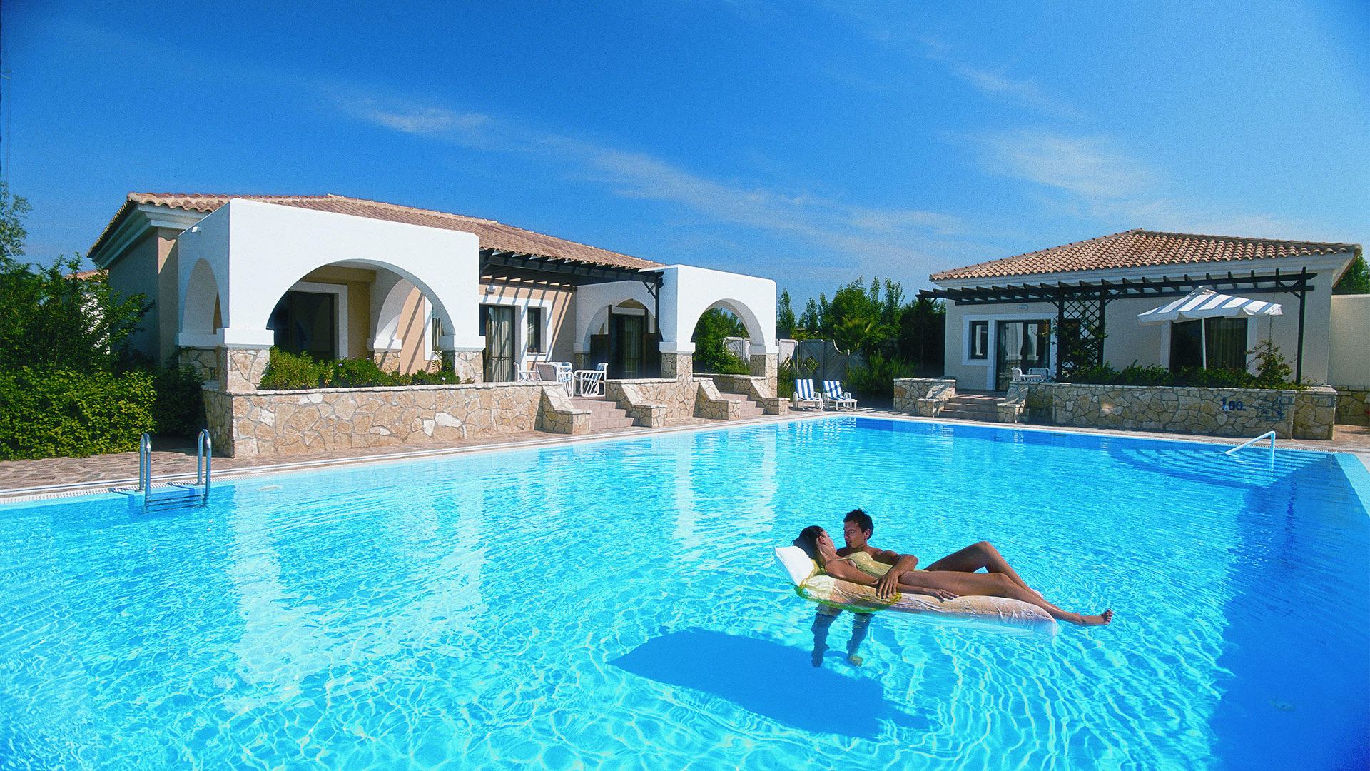 Couple relaxing on a float in a large pool surrounded by white bungalows with arches, tiled roofs and greenery