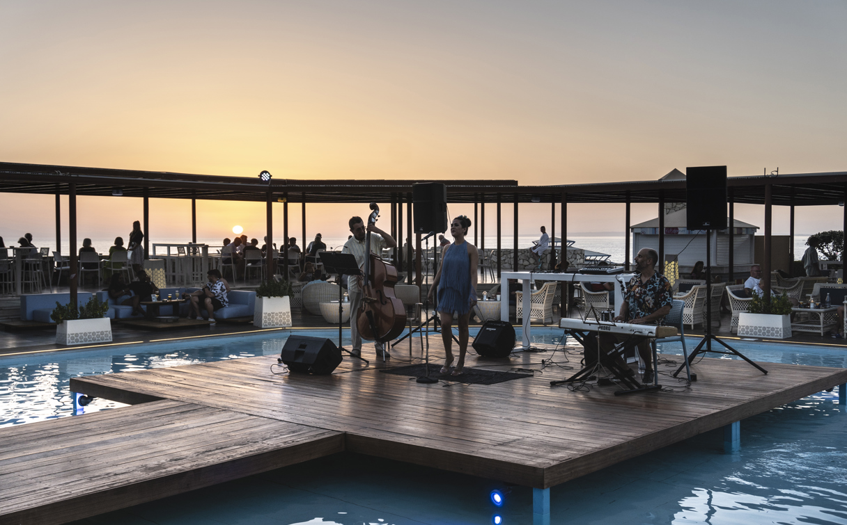 Live band with singer, double bass and keyboard on a wooden stage in the pool, as guests watch the sunset