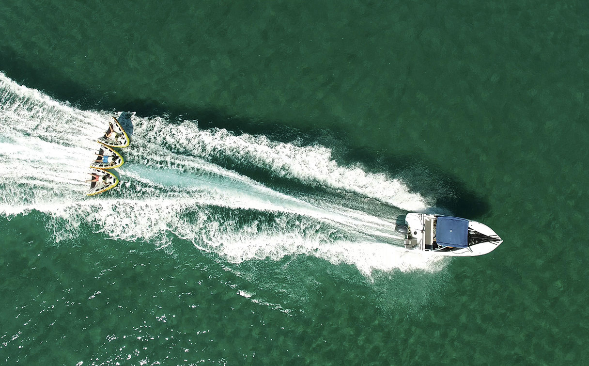 Aerial view of a speedboat towing guests on an inflatable ride, leaving a white wake across the green sea